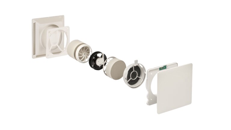 Das Bild zeigt ein in Einzelteile zerlegtes dezentrales Lüftungsgerät. Die Komponenten sind in einer Reihe angeordnet, von der äußeren Abdeckung über den Wärmespeicher, den Ventilator, Filterelemente bis hin zur Innenabdeckung mit Elektronikmodul. Die Darstellung veranschaulicht den Aufbau und die Funktionsweise des Lüftungssystems.